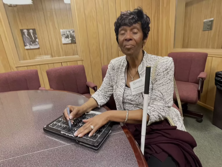 A woman uses a stylus to practice braille on a BrailleDoodle at a table.
