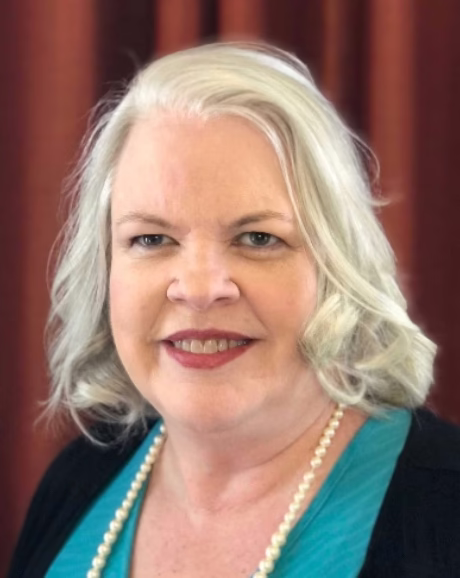 A caucation woman with long gray hair , wearing a black jacket over a teal top smiles broadly.